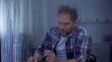 Alkol bağımlısı adam yağmurlu pencerearkasında yalnızlık içinde votka şişesi bakıyor