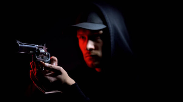 Male thief in hoodie aiming gun against dark background, armed robbery, crime