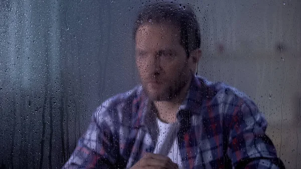Depressed man looking through rainy window, poverty and unemployment concept