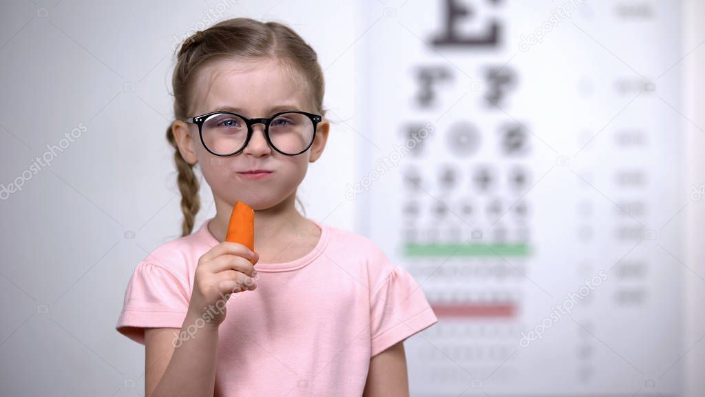 Lovely girl in glasses chewing carrot, vitamin A for good vision, healthcare