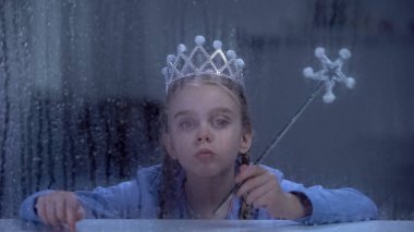 Lonely little princess in crown with magic stick sitting behind rainy window clipart