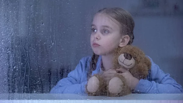 Mała Dziewczynka Przytulanie Misia Patrząc Przez Okno Deszczowej Pogodzie — Zdjęcie stockowe