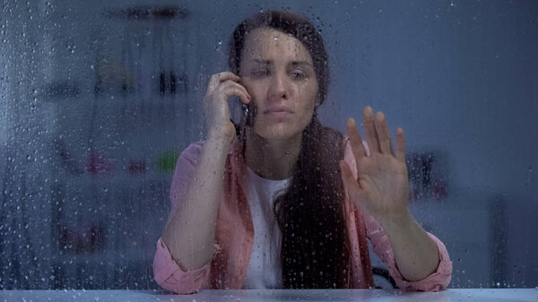 Женщина разговаривает по телефону и смотрит в дождливое окно, ожидая мужа ночью
