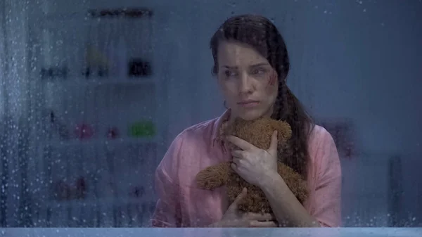 绝望的妇女与受伤的脸拥抱泰迪熊在雨窗后面 — 图库照片