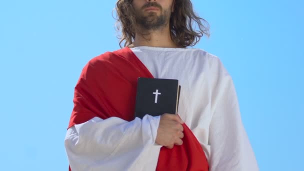 Иисус в мантии и поясе держит святую Библию рядом с сердцем на фоне неба — стоковое видео