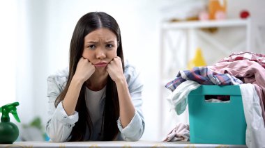 Üzgün ev hanımı kameraya bakıyor, çamaşır sepeti masada kıyafetlerle dolu