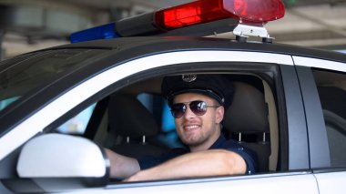 Güneş gözlüğü takan genç polis arabada otururken kameraya gülümseyerek, önemli bir iş