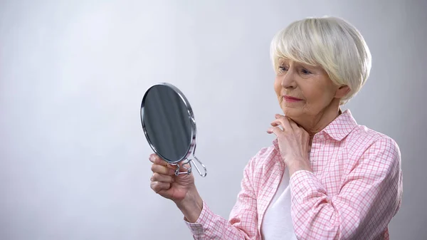 Elderly woman looking at wrinkled face in mirror, thinking about plastic surgery