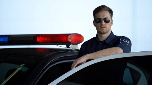 Professional police officer monitoring road standing near door of patrol car