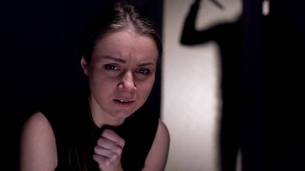 Crying scared woman looking at camera, maniac with knife shadow outside door