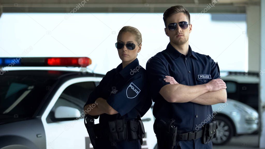 Brave policewoman and man in sunglasses standing with folded hands near car