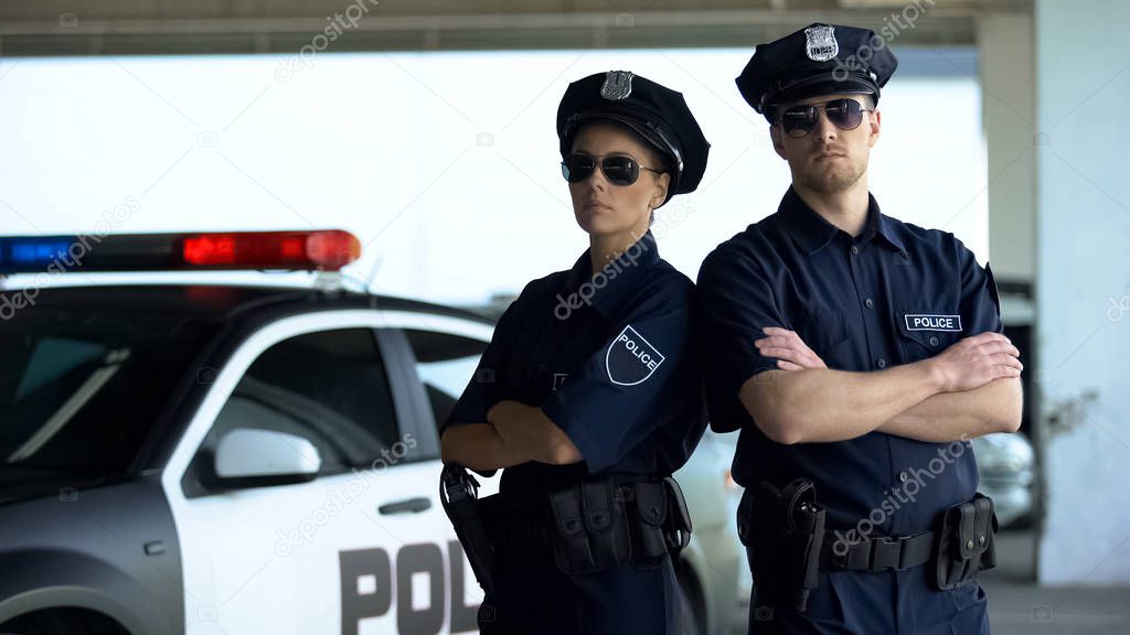 Confident police teammates in uniform and sunglasses standing against patrol car