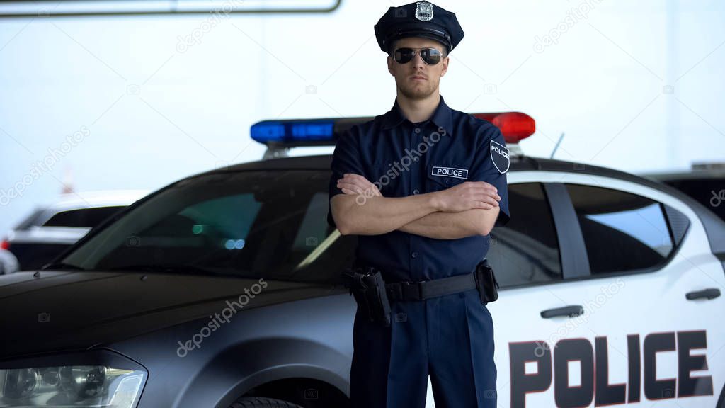 Brave police officer in service cap and sunglasses posing into camera near car