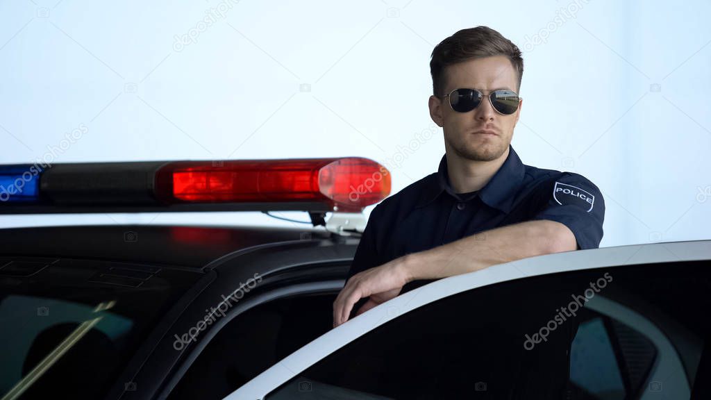 Serious patrolman in sunglasses monitoring road standing near door of police car