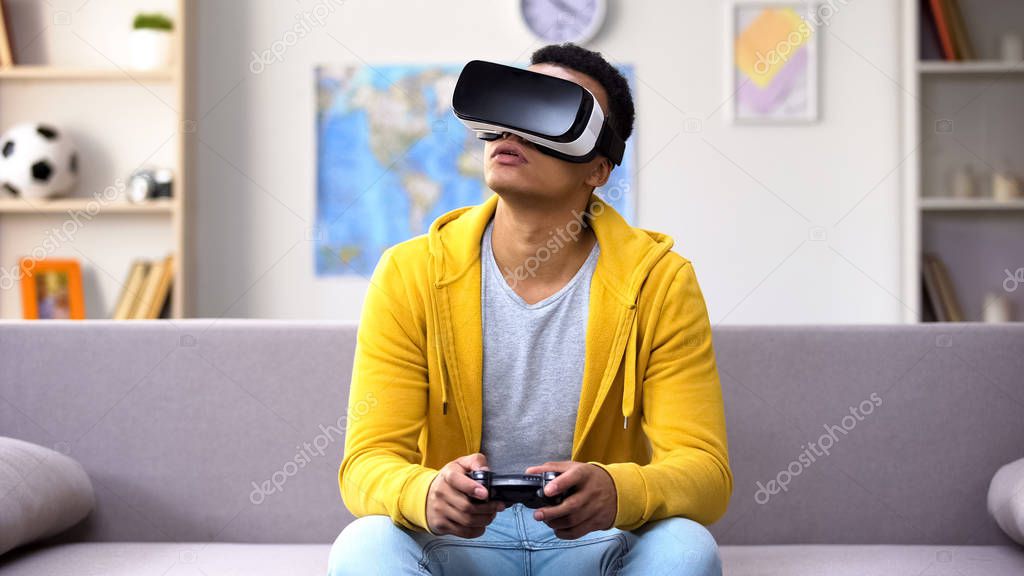 Afro-American teenage boy playing video game in VR headset, modern technologies
