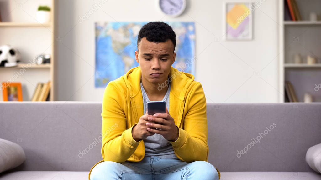 Bored black teenage guy scrolling smartphone, wasting time in social network