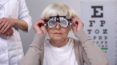 Doctor putting phoropter on elderly woman eyes, choosing proper lens diopter clipart