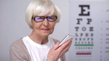 Blind senior woman in eyeglasses holding smartphone and looking at camera clipart