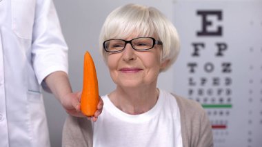 Oculist yaşlı bayan gülümseyen havuç öneren, görme için doğal vitaminler