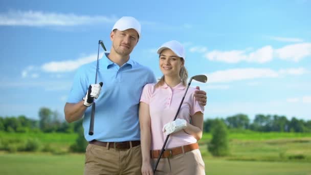 Joven hembra y macho con palos de golf abrazando y sonriendo en cámara, estudio deportivo — Vídeos de Stock