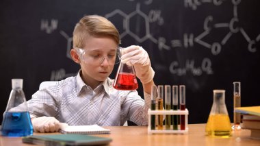 Koruyucu eldiven ve gözlüklü çocuk, kırmızı sıvı ile şişedeki tortuları inceliyor