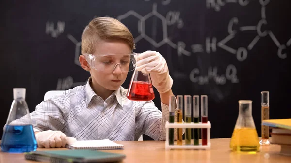 Koruyucu eldiven ve gözlüklü çocuk, kırmızı sıvı ile şişedeki tortuları inceliyor