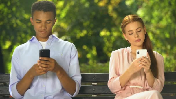 Hombre y mujer adolescentes sentados en el banco y charlando en los teléfonos inteligentes, problemas — Vídeos de Stock