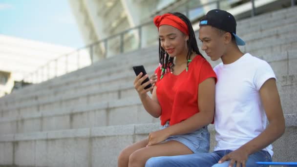 Casal de adolescentes mestiços sentados ao ar livre rolando páginas da web no smartphone — Vídeo de Stock