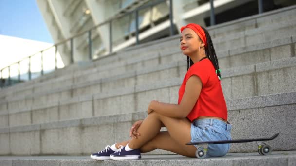 Menina de raça mista atraente com skate descansando ao ar livre, esperando por amigos — Vídeo de Stock