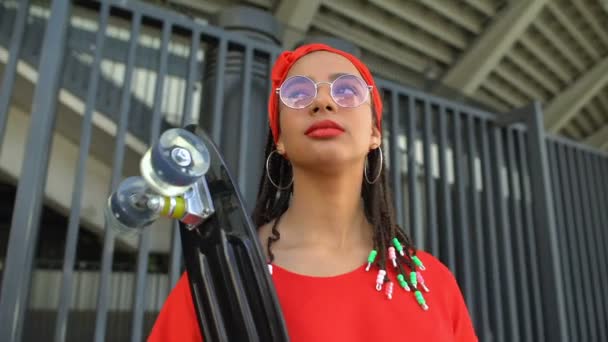 Девушка смешанной расы в модных солнцезащитных очках позирует со скейтбордом, модная фотосессия — стоковое видео