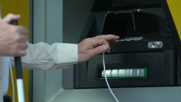 Слепой пожилой человек пытается подключить наушники к банкомату, чтобы снять наличные, банковские — стоковое видео