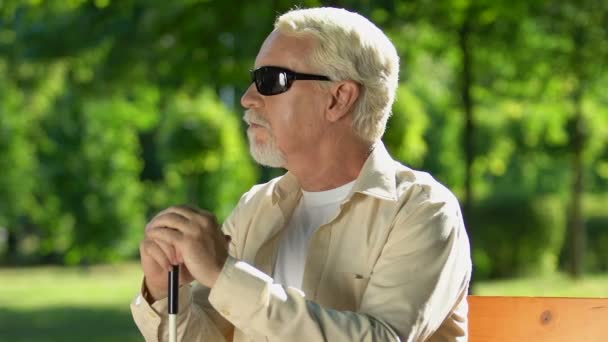 Старший мужчина с проблемами со зрением сидит на скамейке в парке наслаждаясь природой и воздухом — стоковое видео