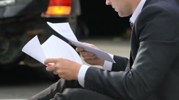 Молодий чоловік читає страховий документ, що сидить на зламаному автомобілі — стокове відео