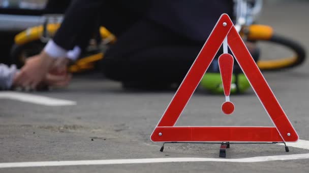 Road danger sign on road, man checking pulse of unconscious bicyclist background — Stock Video