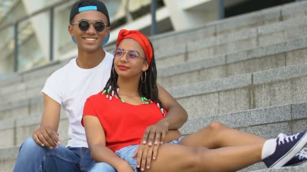 Пара подростков смешанной расы обнимается на ступеньках стадиона, смотрит спортивную игру — стоковое видео