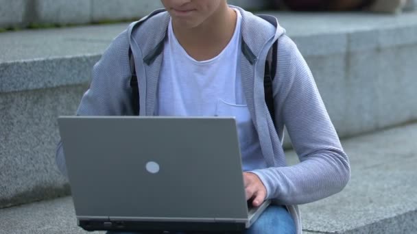 Молодой мужчина, работающий на ноутбуке на улице, это гений или умный подросток-хакер, приложение — стоковое видео