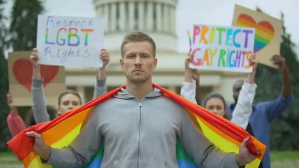 Красивый мужчина с радужным флагом на фоне протестующих за права геев, ЛГБТ-гордость — стоковое видео