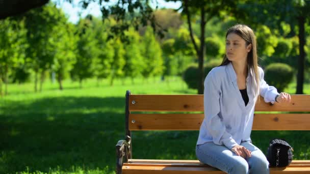 Pacient dívka dívá na hodinky čeká na přítele sedí na lavičce v parku — Stock video