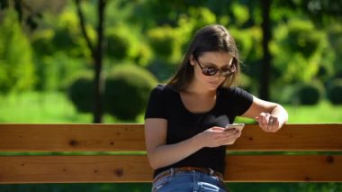 Güneş gözlüklü kız akıllı telefonuyla Sunny Park 'ta bankta dinleniyor, rahatla.