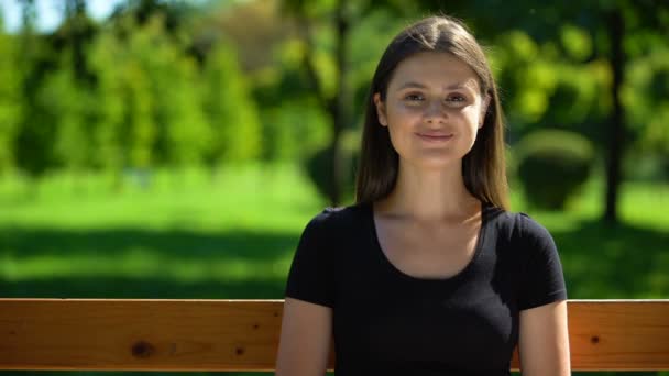 Щаслива молода жінка сидить на лавці в парку і посміхається, дозвілля на відкритому повітрі — стокове відео