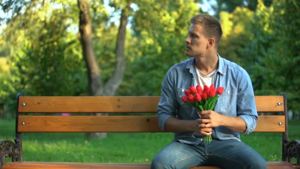 Занепокоєний чоловік з купою тюльпанів чекає дівчину на побачення на лавці в парку — стокове відео
