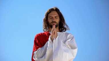 Jesus in robe praying against blue sky background, asking God to forgive sinners clipart
