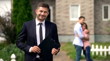 Gülümseyen broker belgeleri ile ayakta, mutlu aile yeni evlerinin yakınında sarılma