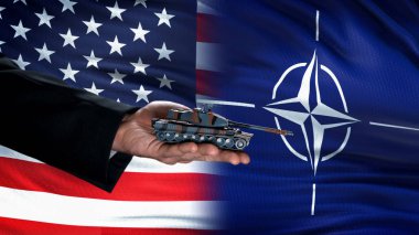 Abd ve Nato bayraklarına karşı oyuncak tankı tutan resmi el, silah ticareti konsepti