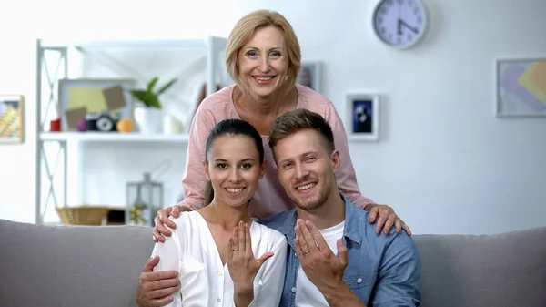 Mother-in-law hugging young couple showing hands with engagement rings at home