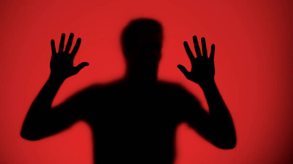 Male silhouette touching glass wall, red lights background, kidnapping victim