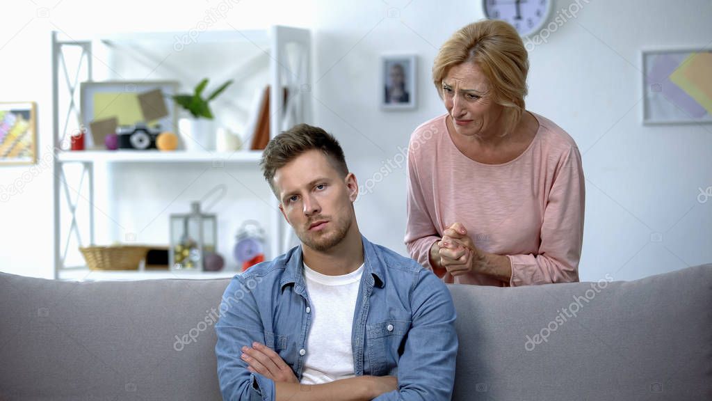 Disappointed mom talking to lazy adult son sitting on sofa at home, upbringing
