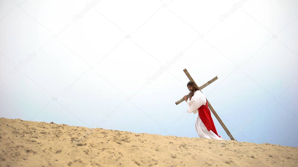 Personification of Jesus Christ carrying heavy cross in desert, crucifixion