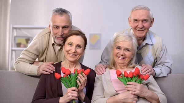 Пожилые Пары Обнимаются Улыбаются Камеру Женщины Держат Тюльпаны Празднуют — стоковое фото