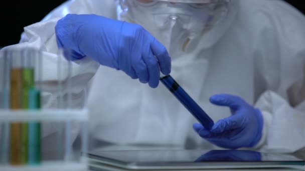 Scientist studying test tube with biohazard liquid and making notes on tablet — Stock Video